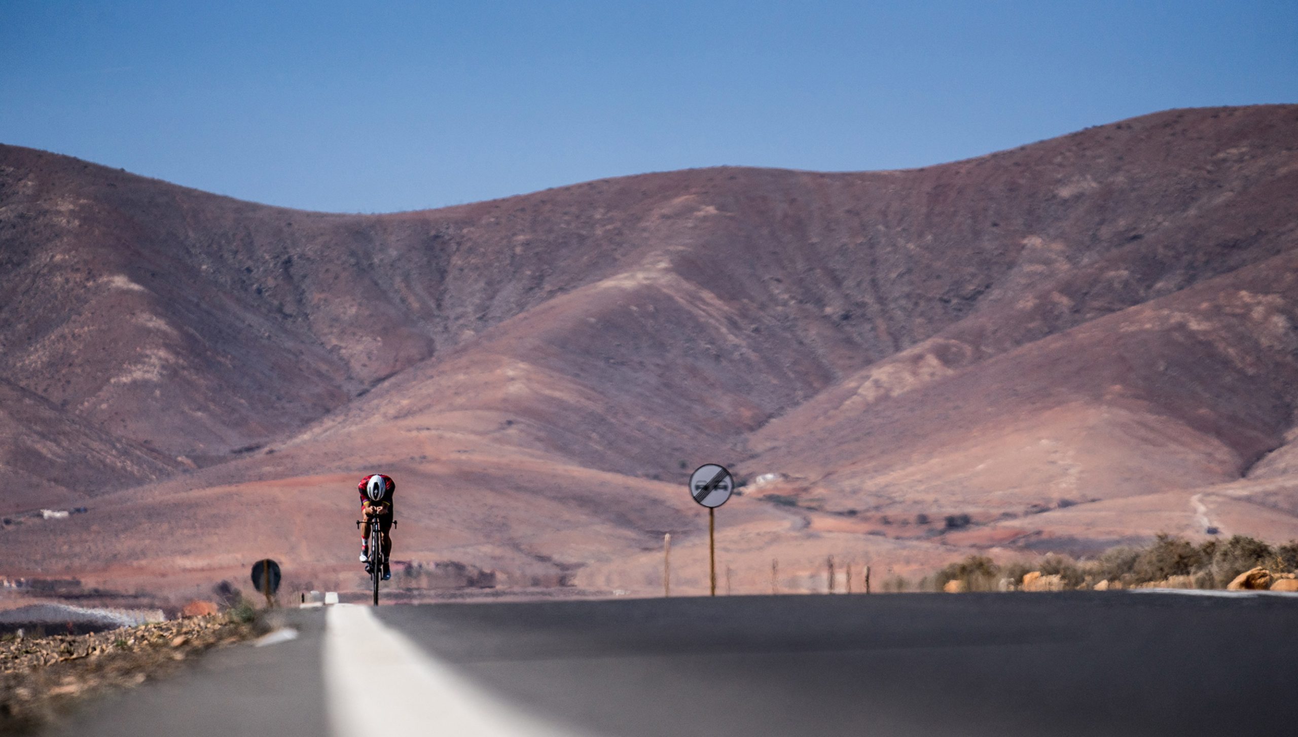 Foto a ras de suelo en una carretera solitaria con un ciclista entrenando de fondo y unas montañas tras él