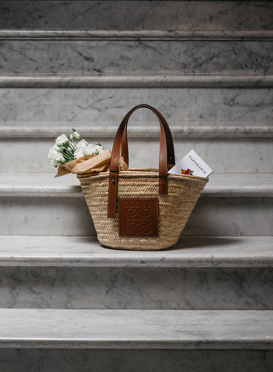 Un bolso de mimbre sobre unas escaleras de mármol blanco con flores blancas y una tableta de chocolates Pancracio