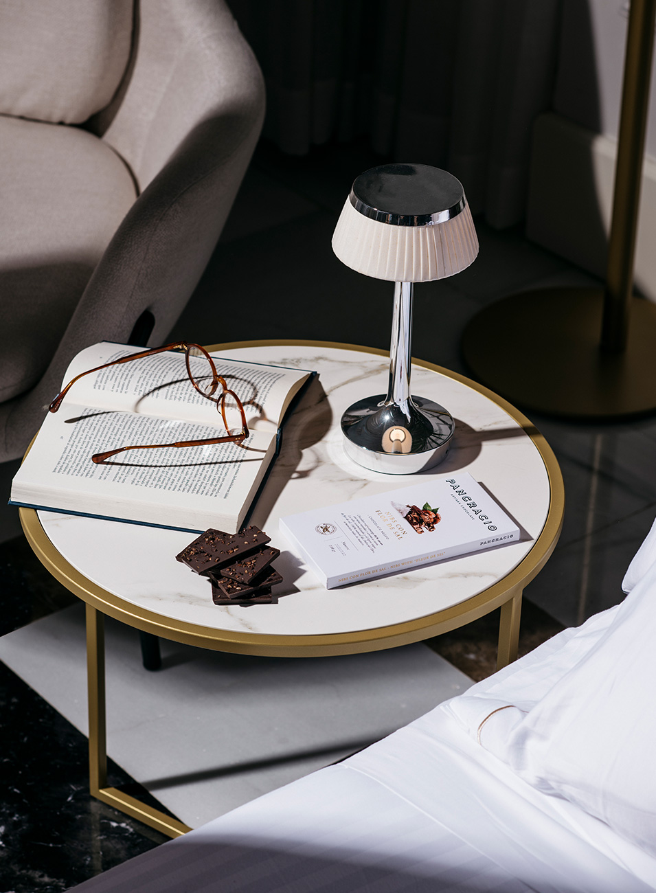 Una mesita baja de salón con una lámpara, unas gafas de leer sobre un libro abierto y una tableta de chocolate con trozos fuera