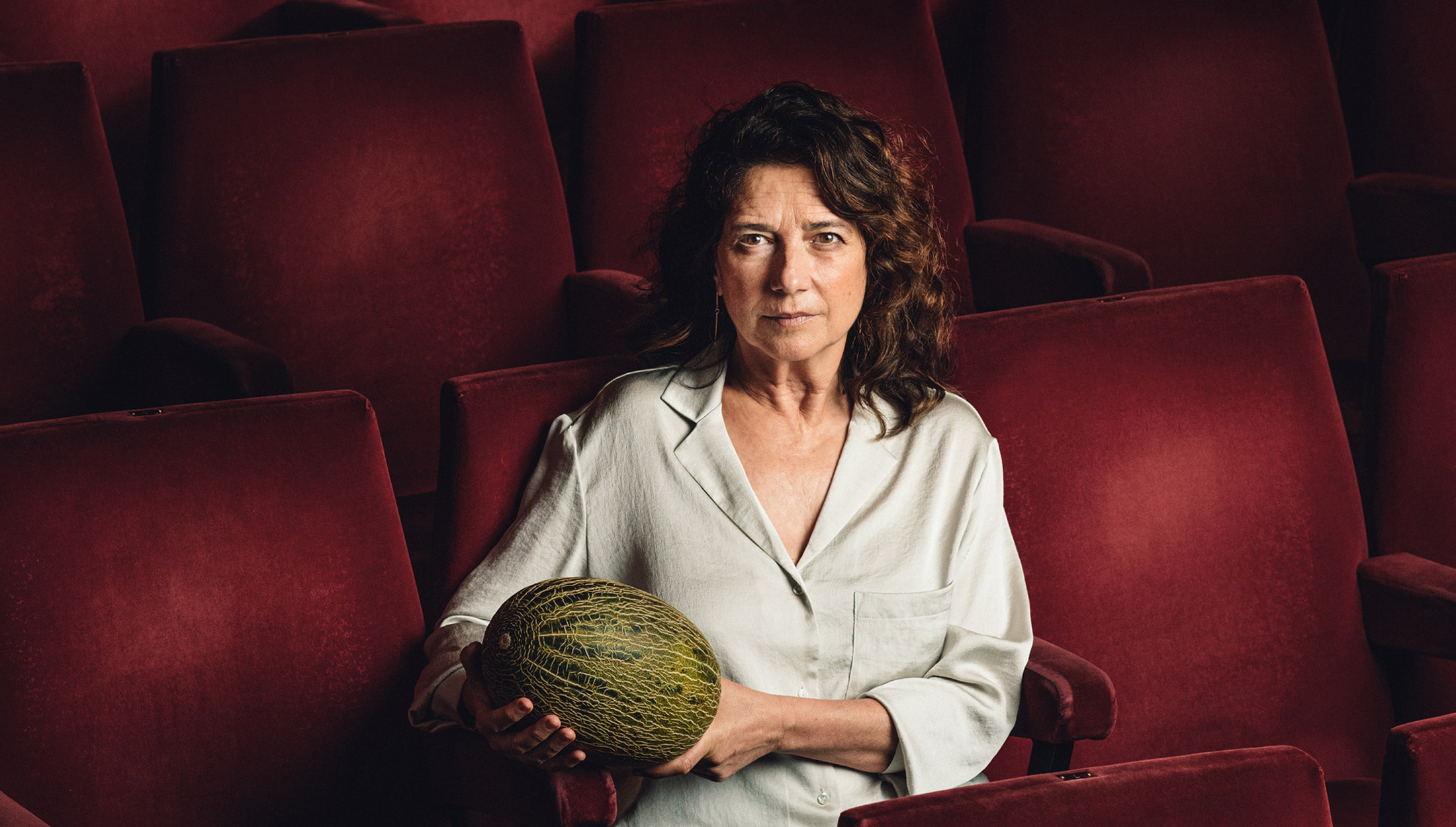 Mujer sentada y sola en una butaca granate de teatro sostiene un melón mirando fijamente a cámara