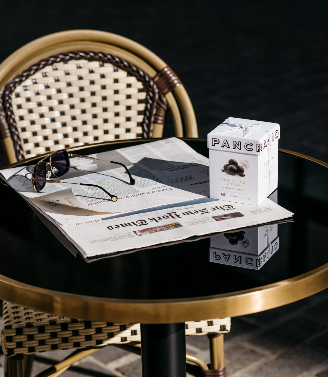 Una mesa al sol, con un periódica y gafas de sol junto a una caja de chocolates Pancracio