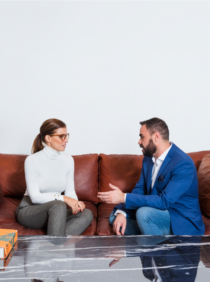 Una mujer y un hombre conversan sentados en un sofá de piel marrón frente a una mesa baja de mármol