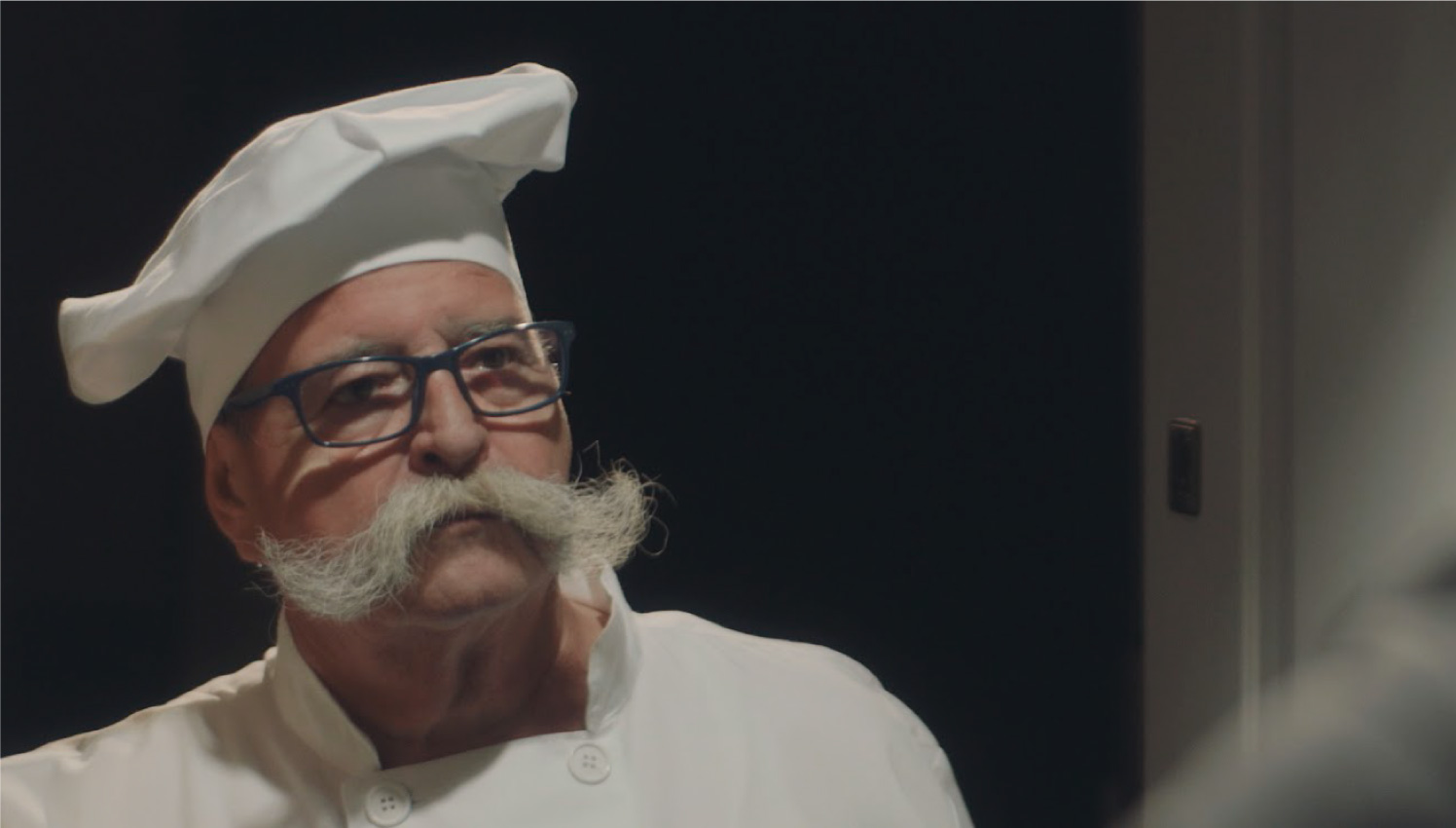 Un chef con sombrero de cocinero, bigote blanco y grande, observa con seriedad