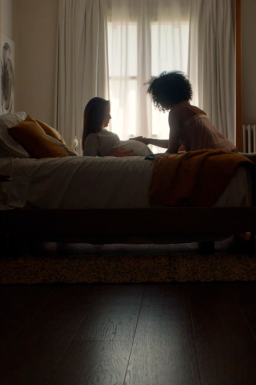 Una foto lejana de 2 mujeres sentadas en la cama, una acaricia la barriga de la otra embrazada