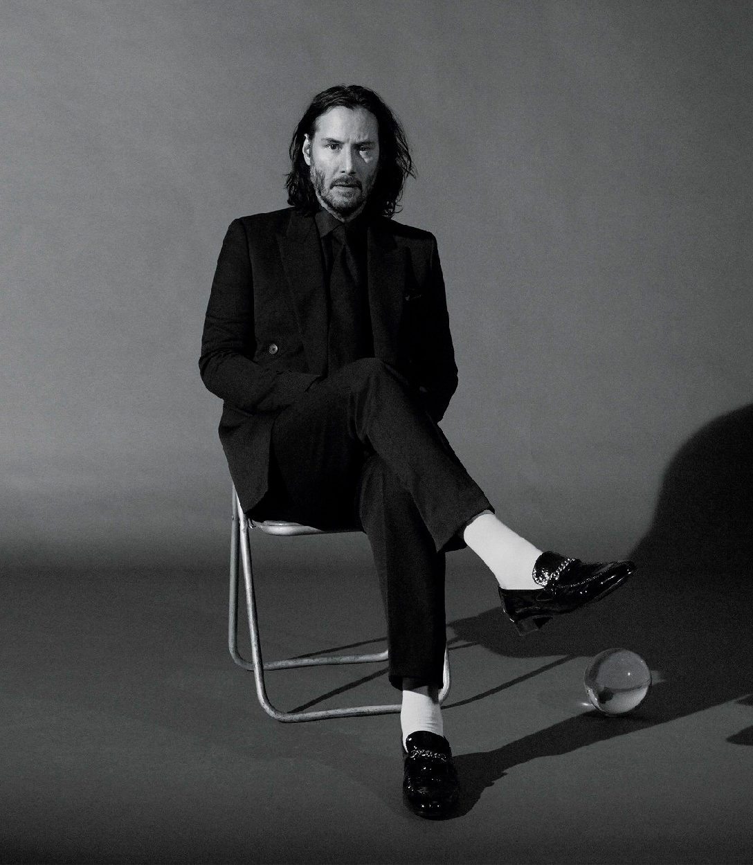 Fotografía de Keanu Reeves con barba sentado en una silla, vestido negro de chaqueta y con calcetines blancos