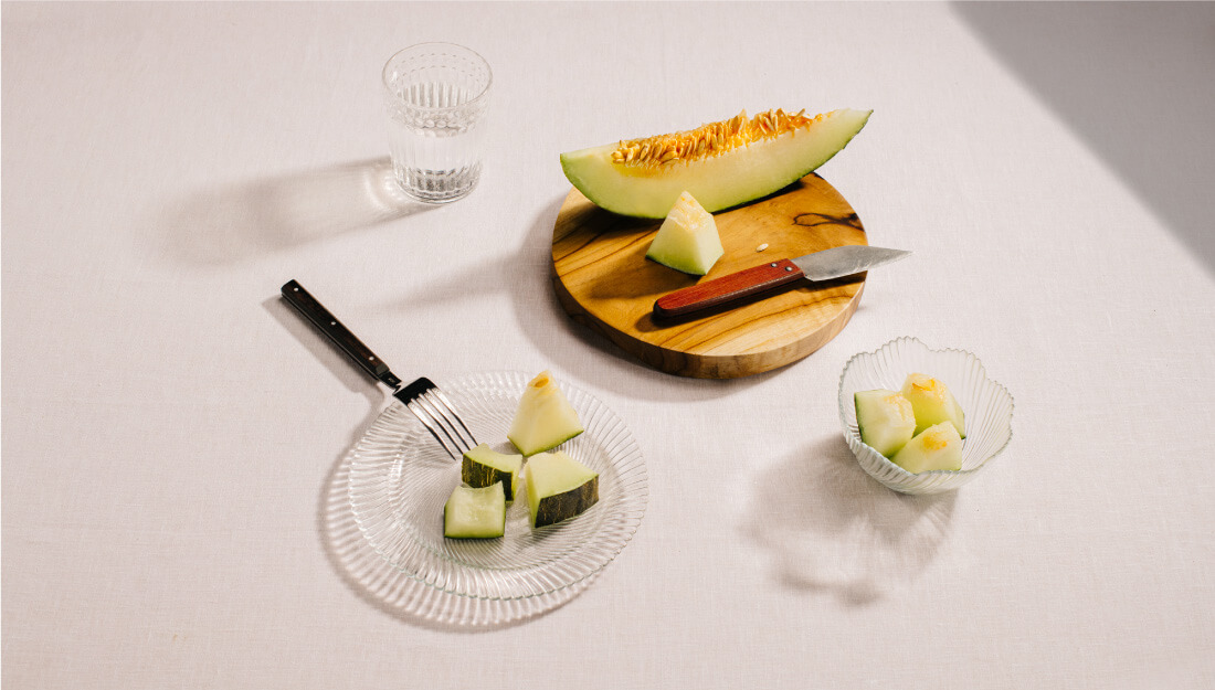 Sobre un mantel blanco, una tabla de madera con un melón cortado, un plato con trozos de melón, una vaso de agua y una tarrina de cristal con más trozos de melón