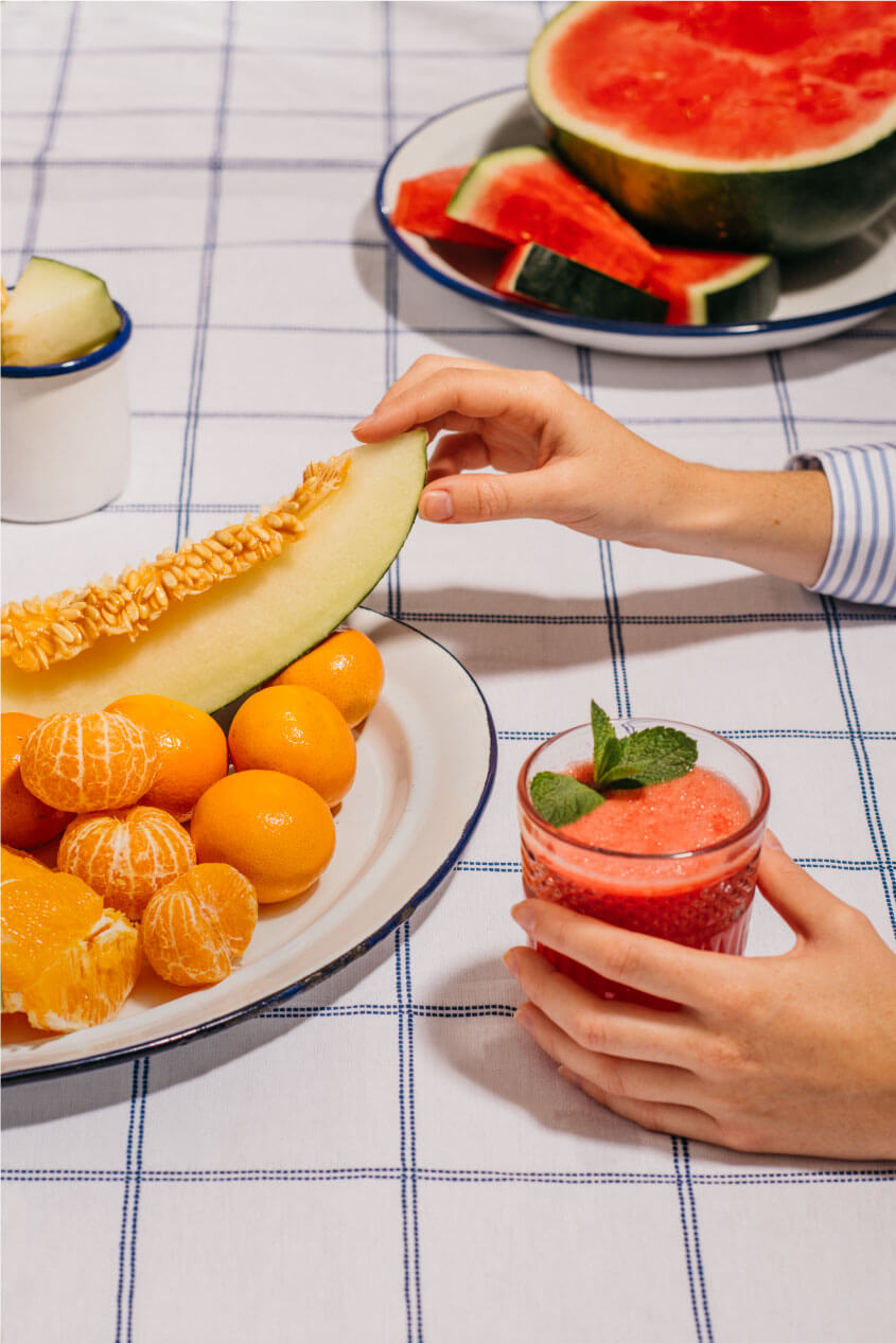 Una mesa llena de frutas con melón, mandarinas y sandía, mientras unas manos cogen un trozo de melón y tiene un zumo rojo en la otra mano