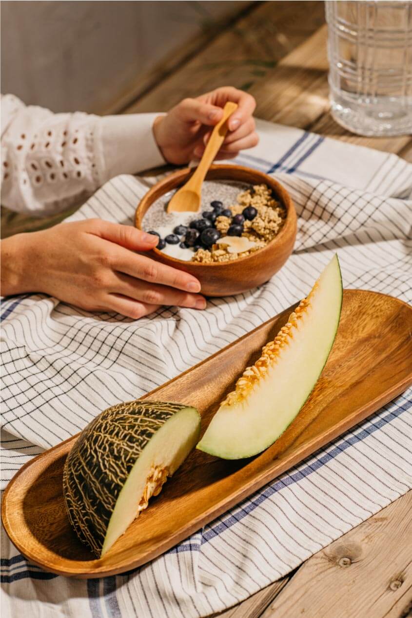 Un melón cortado en una bandeja de madera, mientras alguien desayuna una crema con frutos secos y cerezas