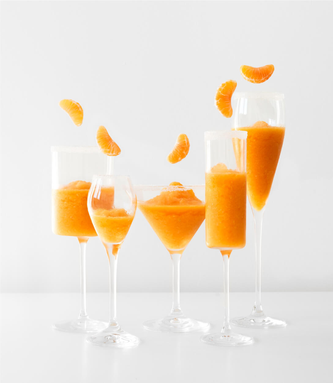 Bodegón de 5 copas con zumo de mandarina mientras al vuelo caen gajos de mandarina