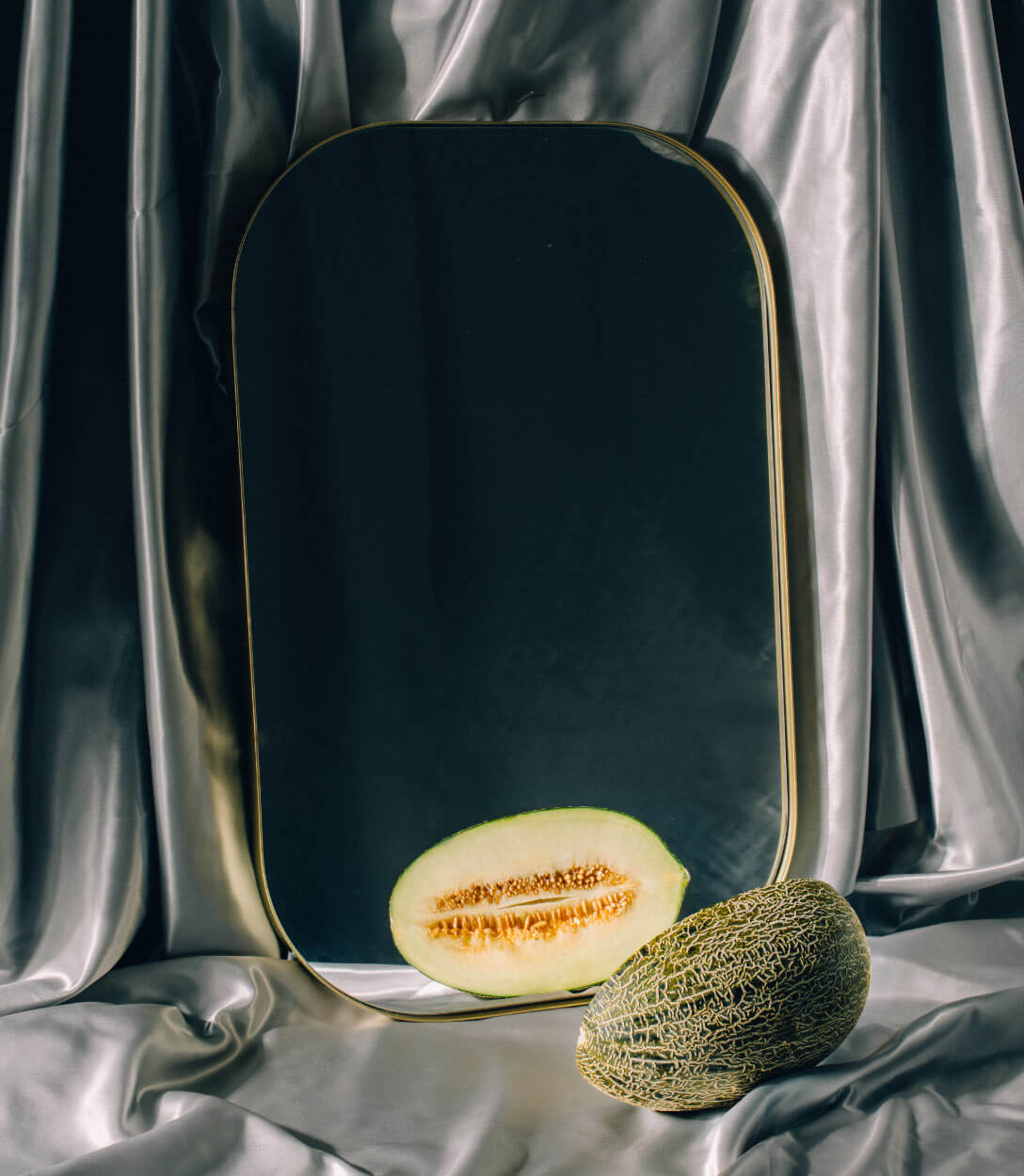 Medio melón frente a un espejo, con unos fondos de tela plateada
