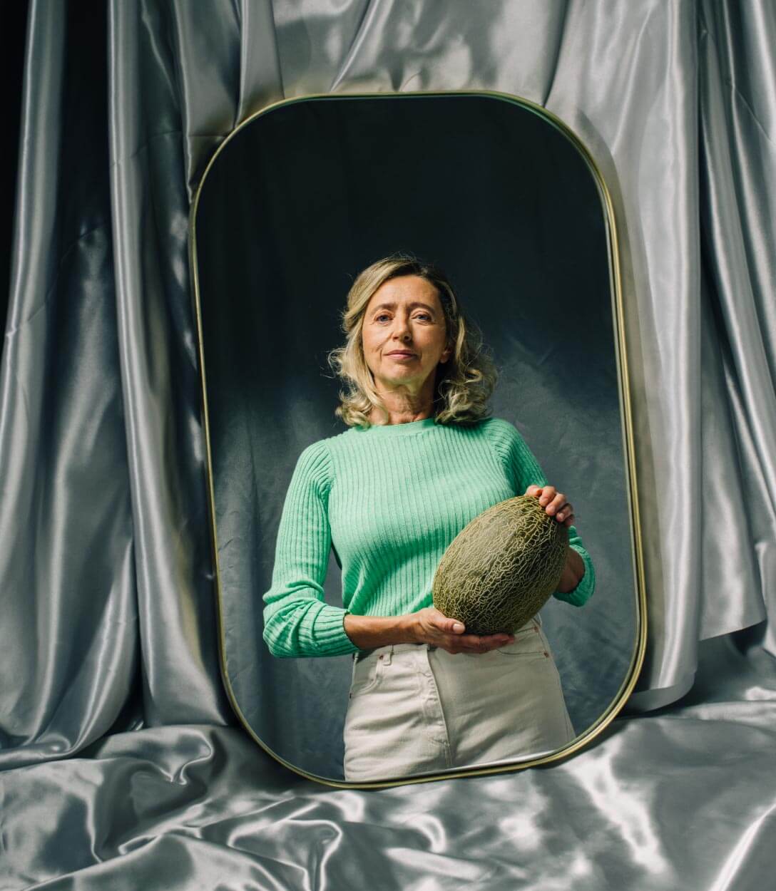 Mujer con suetere verde y pantalón claro se le ve en un espejo sosteniendo un melón
