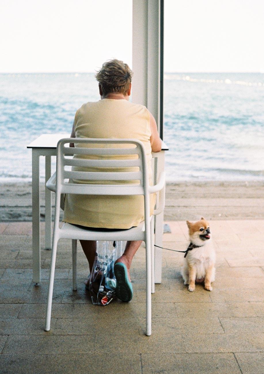 Una mujer sentada en una terraza de la playa de espaldas con su perro pequeño cogido a la correa