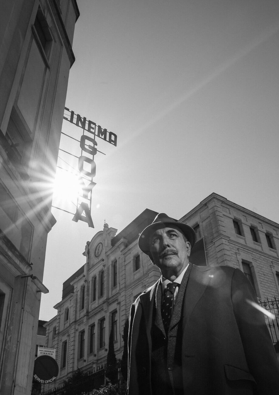Un hombre con gabardina y sombrero mira a su izquierda cerca de los cines Goya