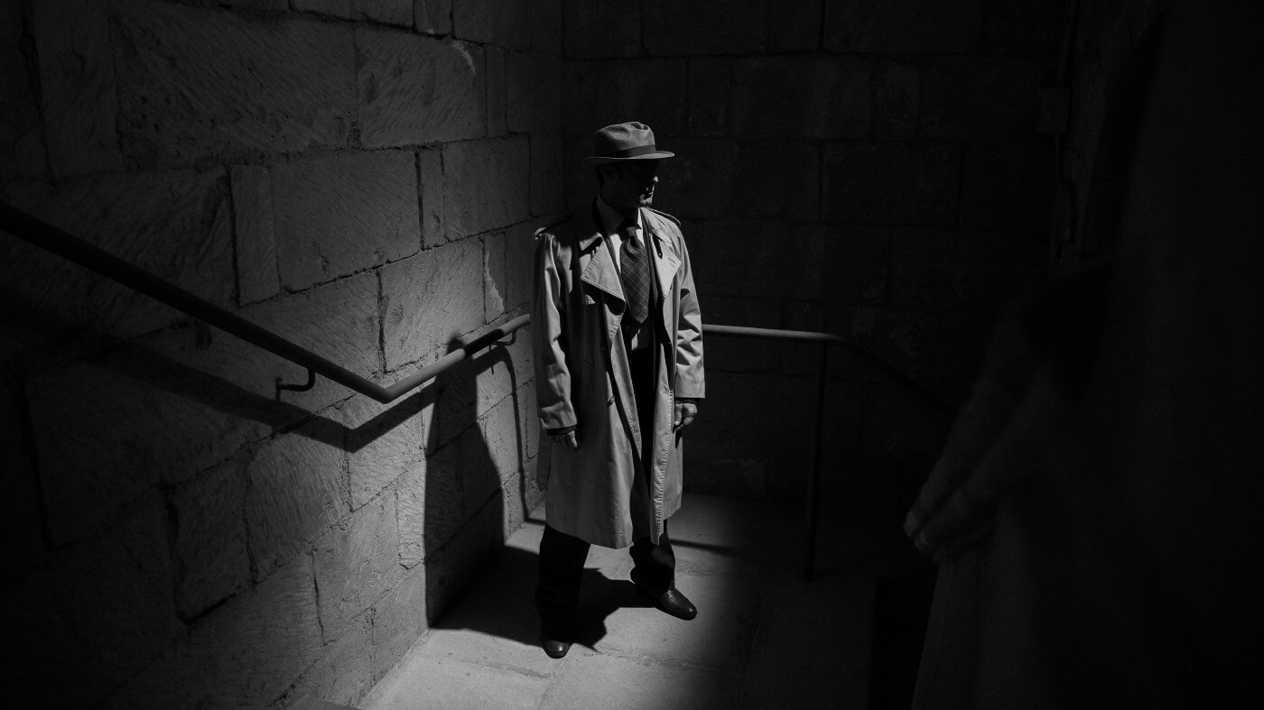 Un hombre con gabardina y sombrero esperando en las escaleras de un edificio con actitud seria