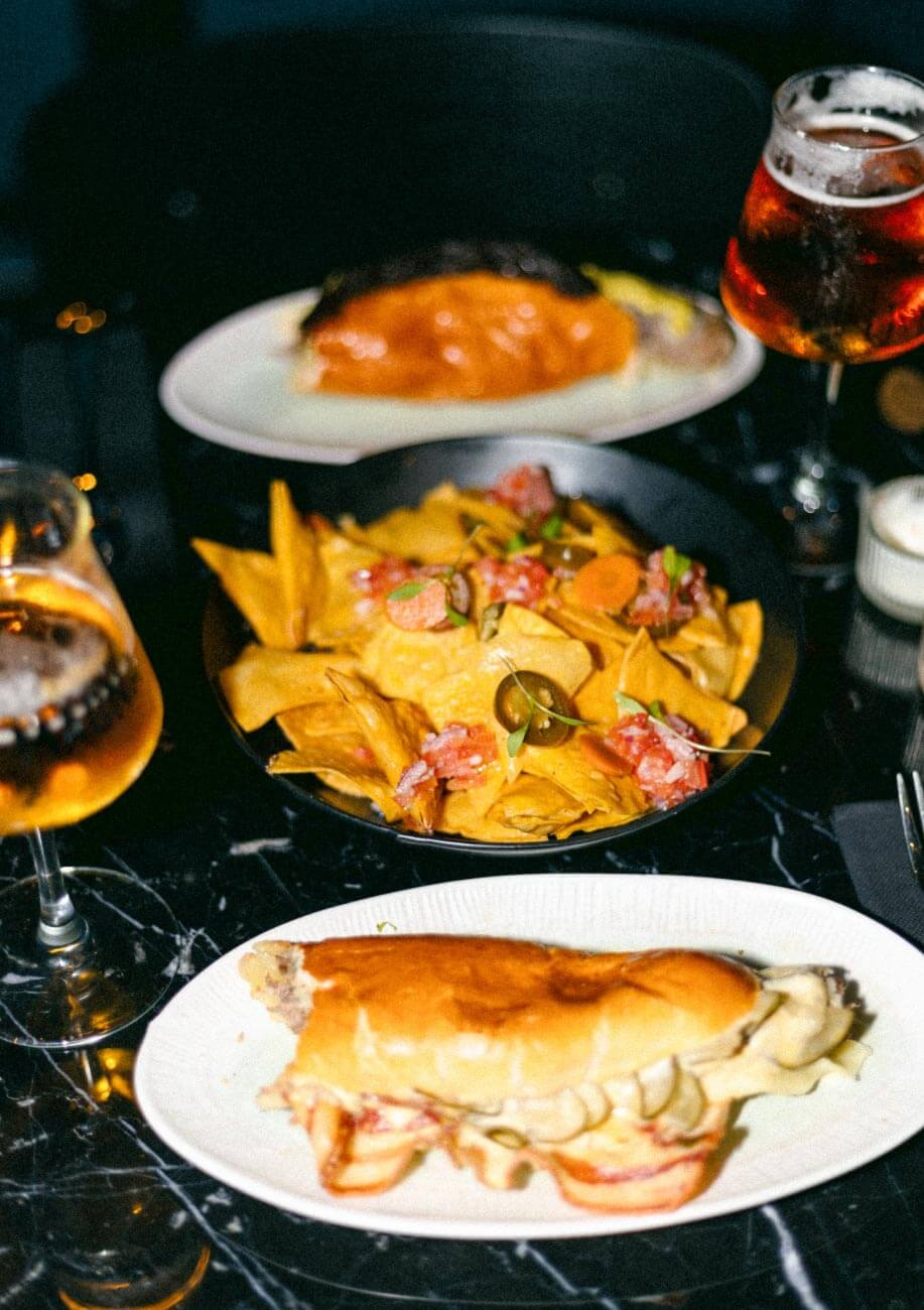 Vista de 3 platos de comida sobre una mesa oscura, cervezas y mesa de mármol negra