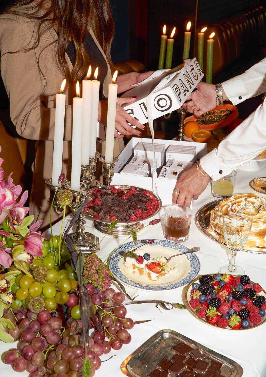 Un hombre y una mujer abren una caja con varios productos de Pancracio sobre una mesa con frutas, velas, flores y varias bebidas