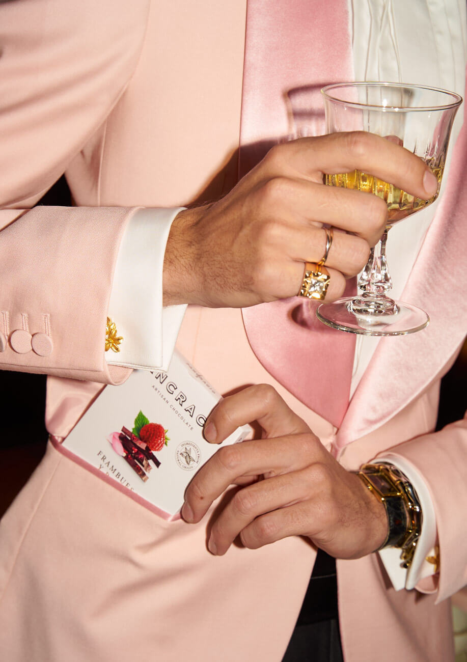 Un hombre saca de su chaqueta rosa una tableta de chocolates Pancracio y en la otra mano una copa de champagne