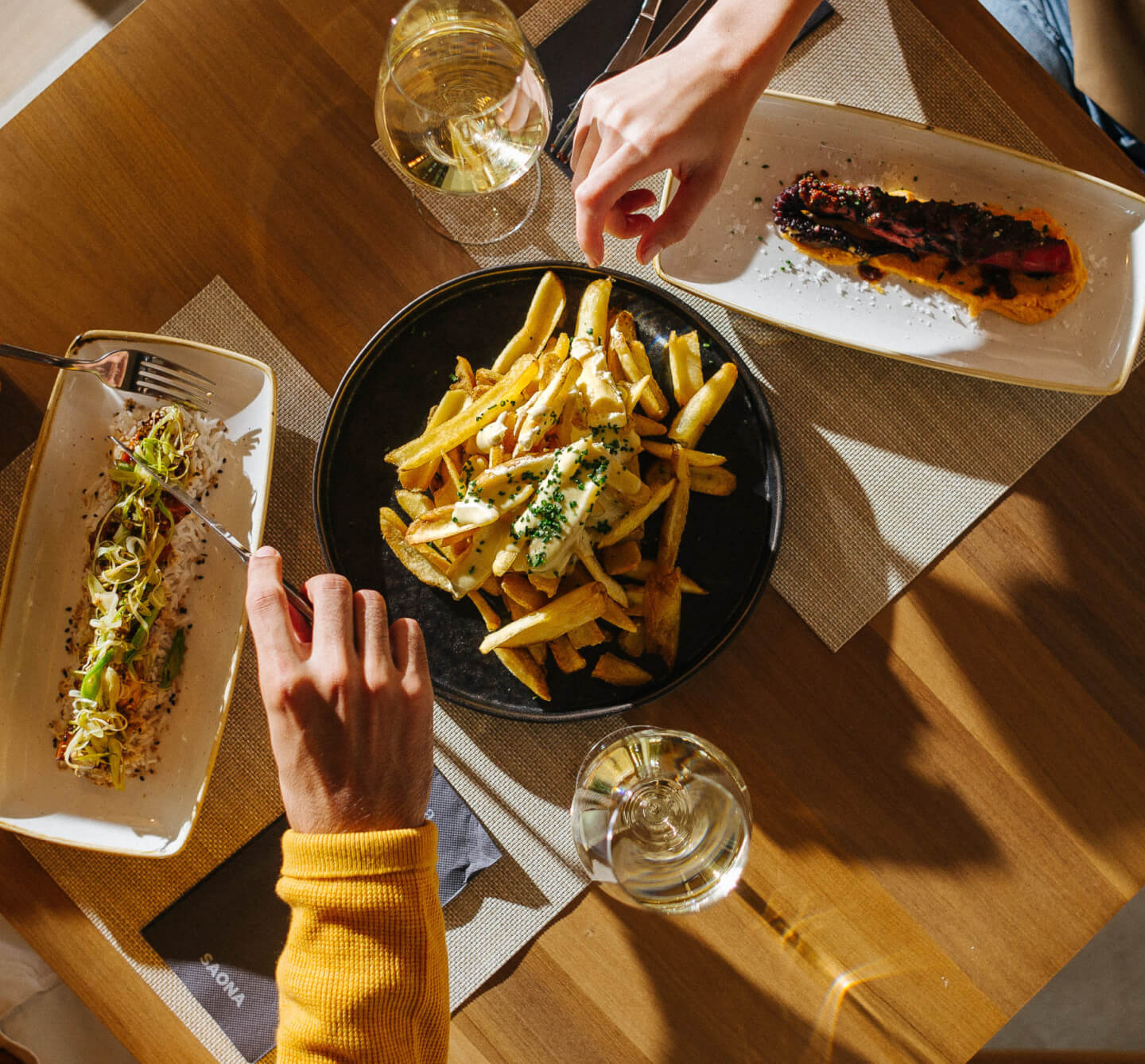 Fotos de 2 brazos cogiend comida en una mesa con dos copas de vino blanco y una luz de atardecer con sombras muy pronunciadas